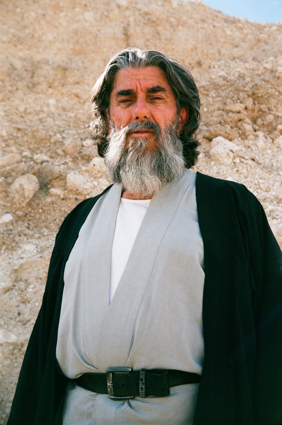 Portrait of a grey-bearded man in robes against a sandstone wall